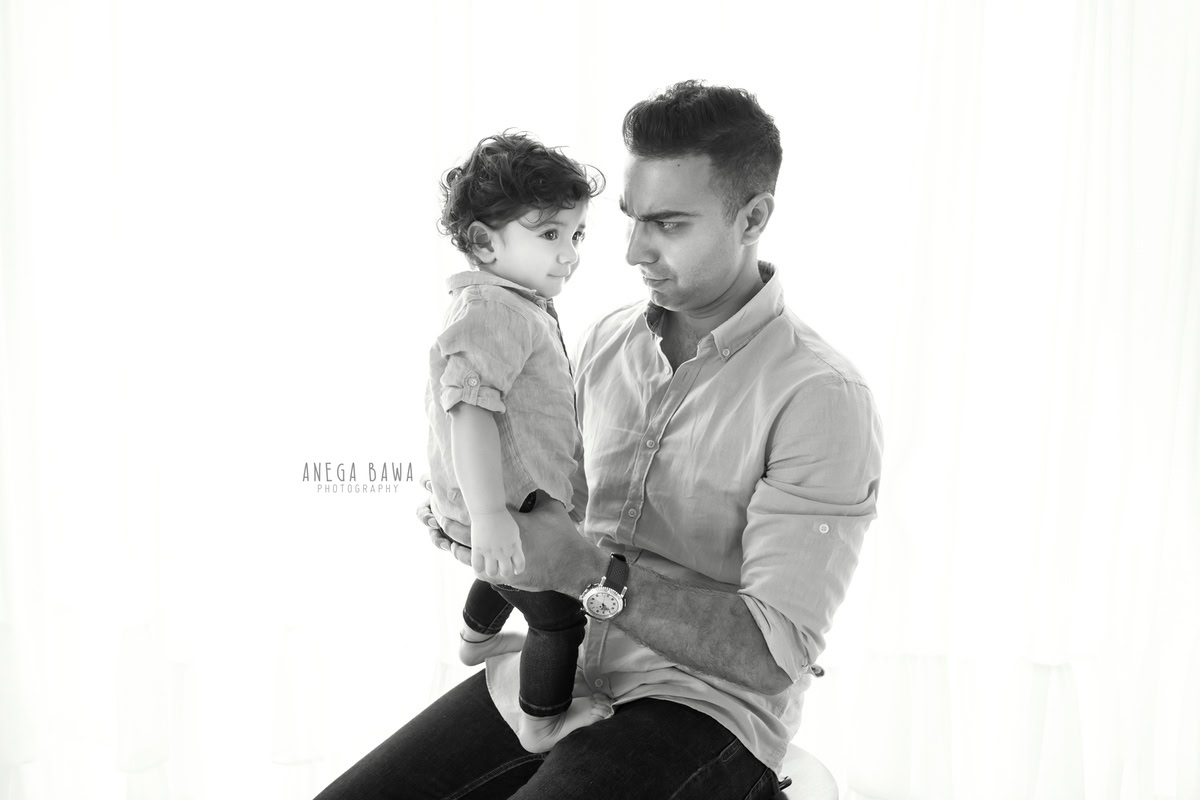 1-year-old boy standing on dad's lap against a white backdrop, captured by Anega Bawa, family photographer in Gurgaon (Delhi NCR).