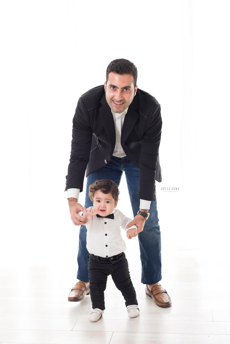 1-year-old boy standing on the floor, holding dad's hands against a white backdrop, wearing a cute bow and white shirt, captured by Anega Bawa, family photographer in Gurgaon (Delhi NCR).