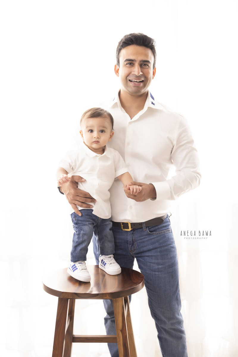 1-year-old boy standing on a stool, posing with dad in a white shirt and denims against a white backdrop, captured by Anega Bawa Photography for a family photoshoot in Gurgaon (Delhi NCR).