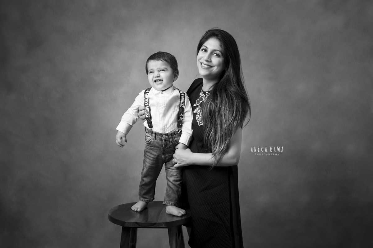 1-year-old boy standing on a stool, posing with mom in a black and white pose against a dull backdrop, captured by Anega Bawa Photography for a family photoshoot in Gurgaon (Delhi NCR).
