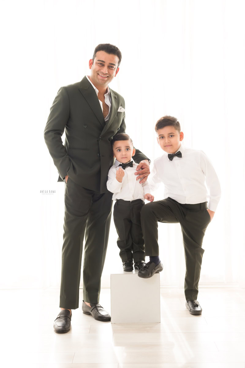 1-year-old boy standing on a white stool, twinning with his elder brother in a white shirt and bow tie, with dad holding his son against a white backdrop, captured by Anega Bawa Family Photographer Gurgaon (Delhi NCR).