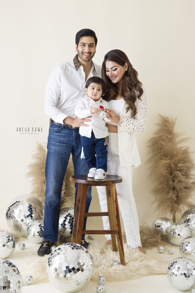 1-year-old boy standing on a wooden stool, surrounded by bushes and disco balls against a beige backdrop."
