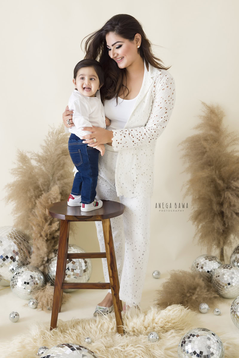 1-year-old boy standing on a wooden stool, posing with mom against a beige backdrop with bushes and disco balls, captured by Anega Bawa Photography for a family photoshoot in Gurgaon (Delhi NCR).