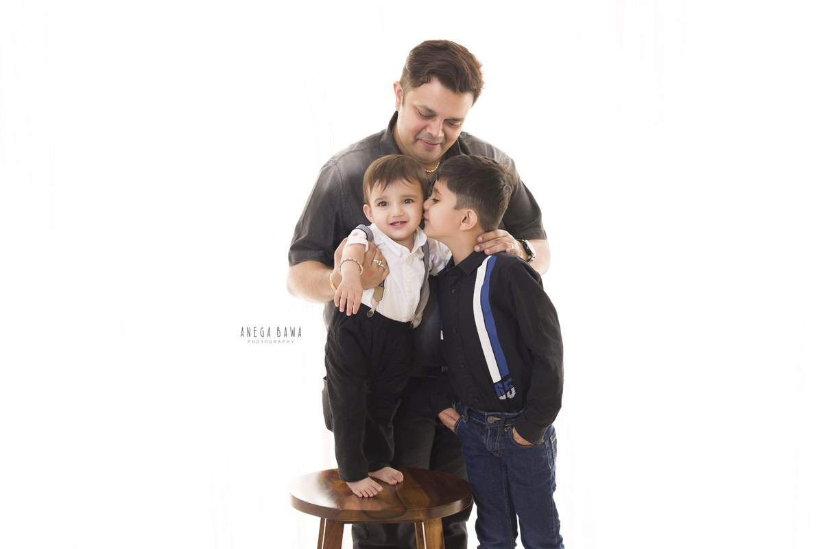 1-year-old boy standing on a wooden stool, with his elder brother kissing dad on the back, against a white backdrop, captured by Anega Bawa Family Photographer Gurgaon (Delhi NCR).