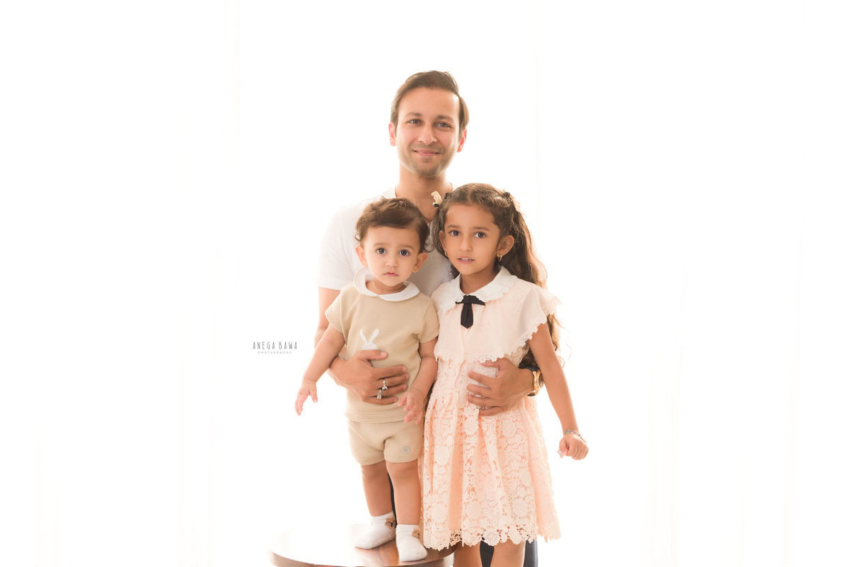 1-year-old boy standing on a wooden stool, posing with dad and elder sister against a white backdrop, captured by Anega Bawa, family photographer in Gurgaon (Delhi NCR).