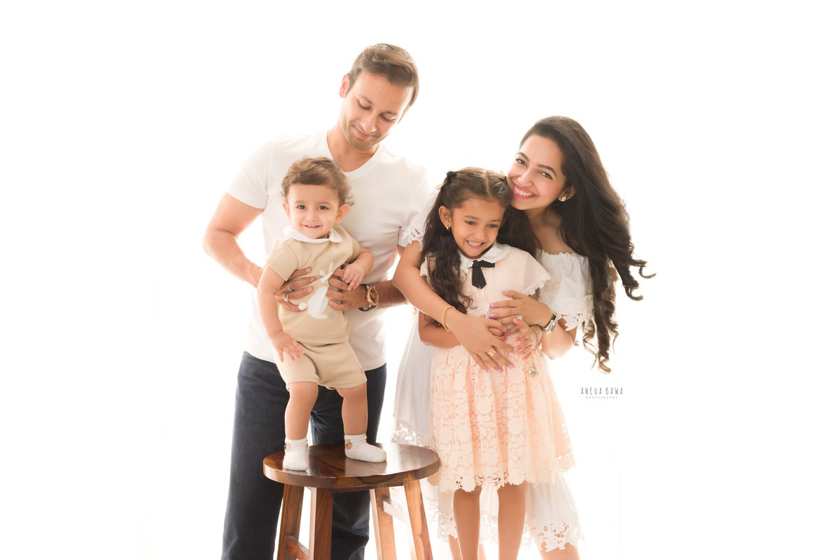 1-year-old boy standing on a wooden stool, posing with dad and elder sister against a white backdrop, captured by Anega Bawa, family photographer in Gurgaon (Delhi NCR).