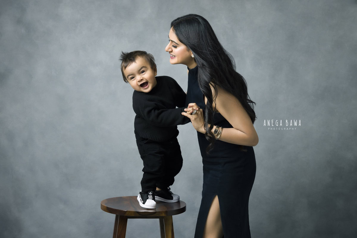 1-year-old boy standing on a wooden stool, posing with mom against a black-grey backdrop, captured by Anega Bawa, family photographer in Gurgaon (Delhi NCR).