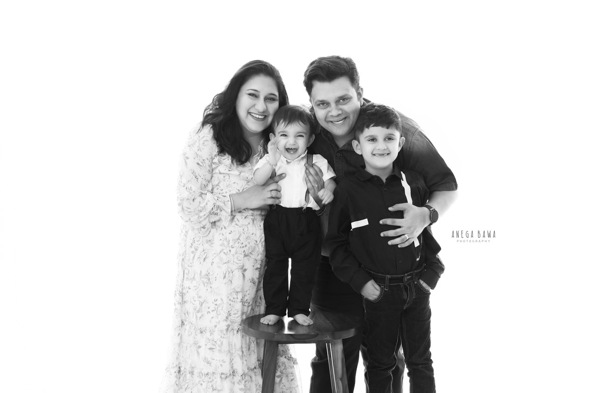 1-year-old boy standing on a wooden stool, posing with mom, dad, and elder brother against a white backdrop, captured by Anega Bawa, family photographer in Gurgaon (Delhi NCR).
