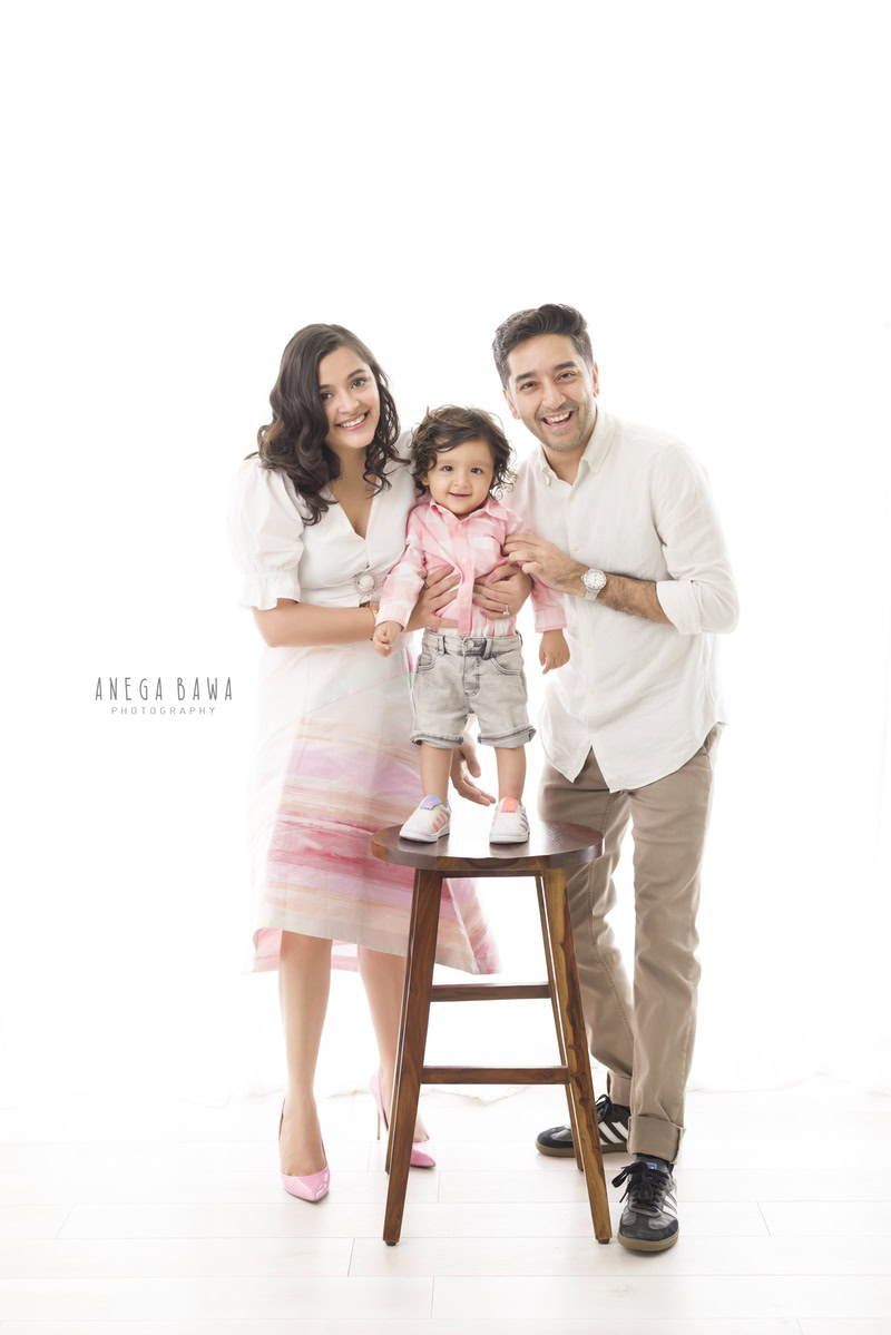 1-year-old boy standing on a wooden stool, posing with mom and dad against a white backdrop, captured by Anega Bawa Family Photographer Gurgaon (Delhi NCR).