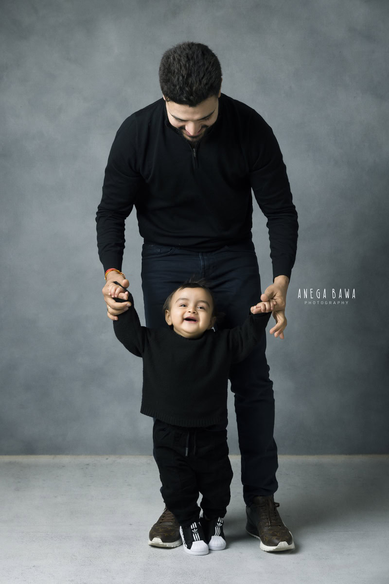 1-year-old boy standing with dad, both twinning in black, against a grey backdrop, captured by Anega Bawa Photography for a family photoshoot in Gurgaon (Delhi NCR)