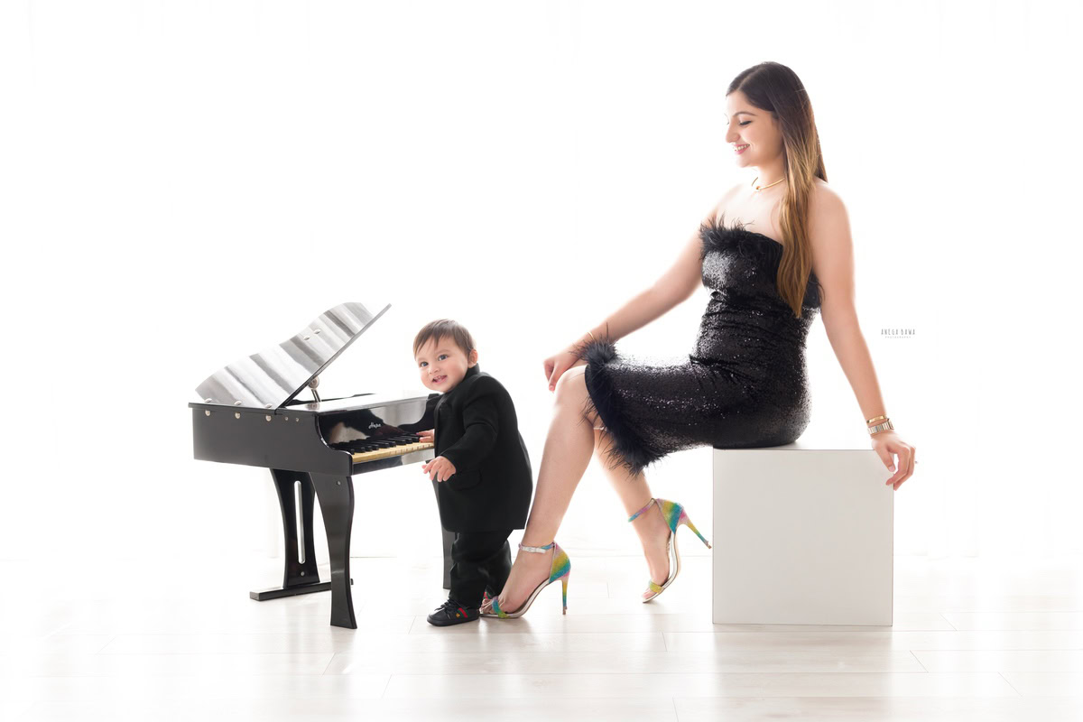 1-year-old boy wearing a black suit, posing with a piano, mom seated on a stool against a white backdrop, captured by Anega Bawa, family photographer in Gurgaon (Delhi NCR).