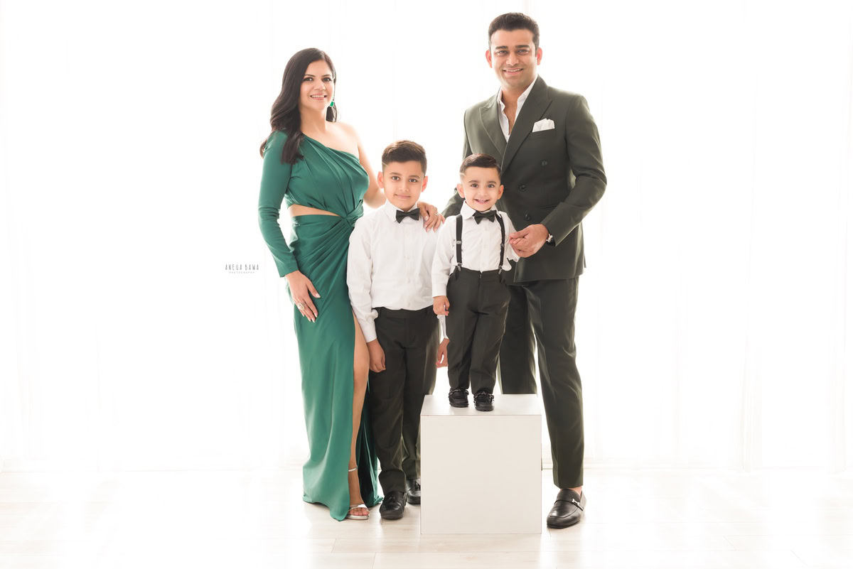 1-year-old boy wearing a cute bow and white shirt, standing on a stool, posing with mom, dad, and elder brother in a family pose against a white backdrop, captured by Anega Bawa Family Photographer Gurgaon (Delhi NCR).