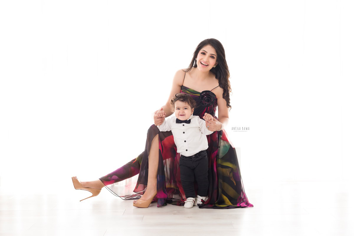 1-year-old boy in a white shirt and bow, posing cutely with mom against a white backdrop, captured by Anega Bawa Photography for a family photoshoot in Gurgaon (Delhi NCR).