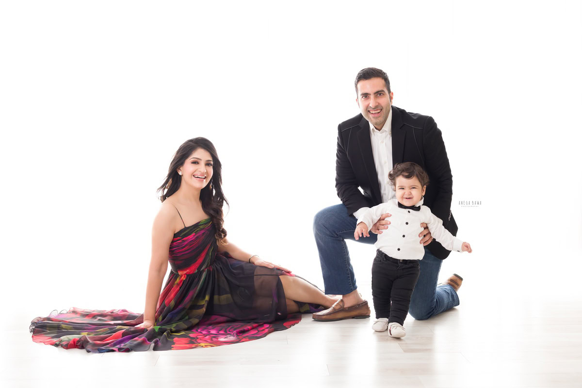 1-year-old boy in a white shirt and bow standing on the floor, posing with dad and mom against a white backdrop, captured by Anega Bawa Family Photographer Gurgaon (Delhi NCR).