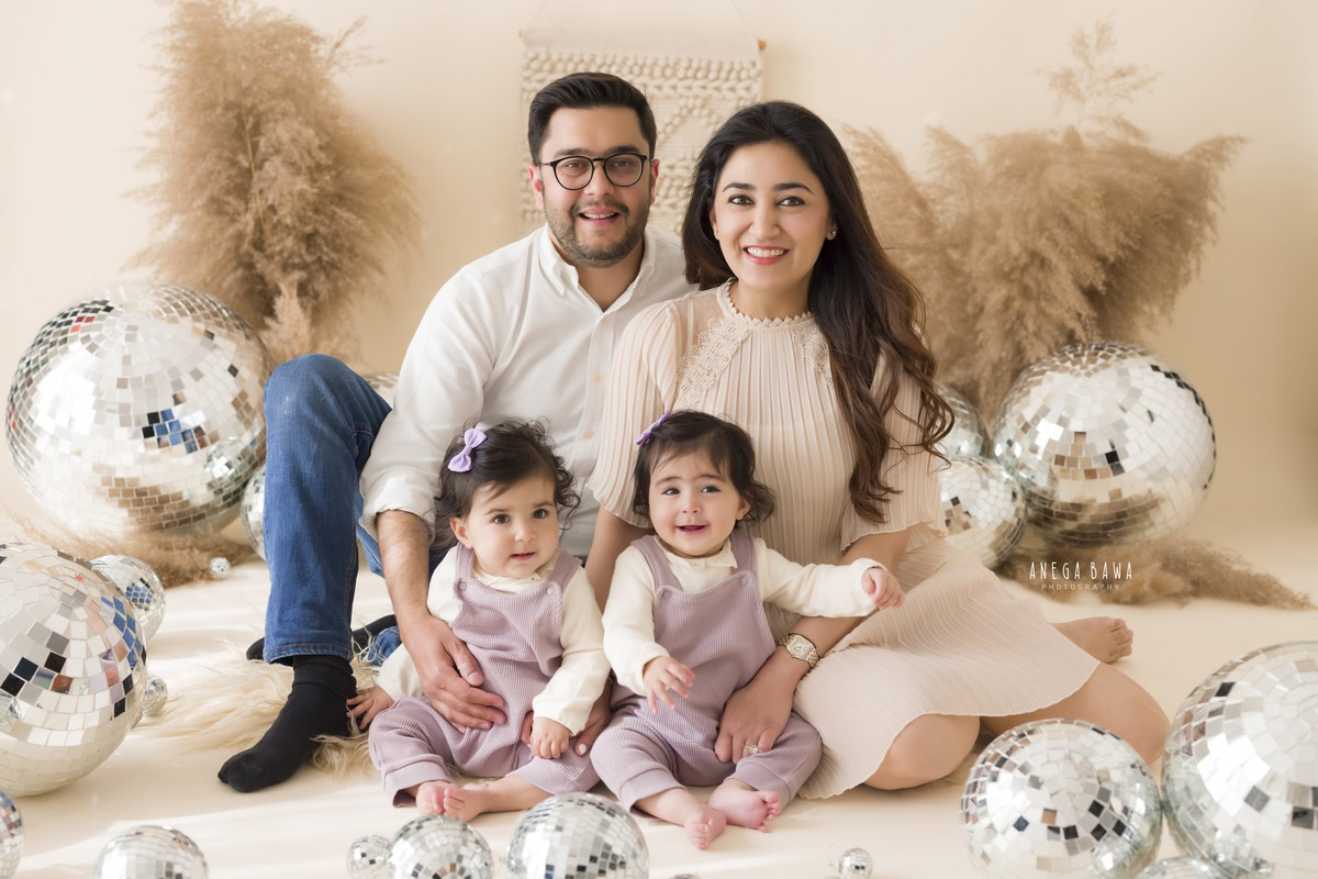 1-year-old cute twins posing with mom and dad amidst bushes and disco balls on a beige backdrop, photography done by Anega Bawa, Gurgaon (Delhi, NCR)