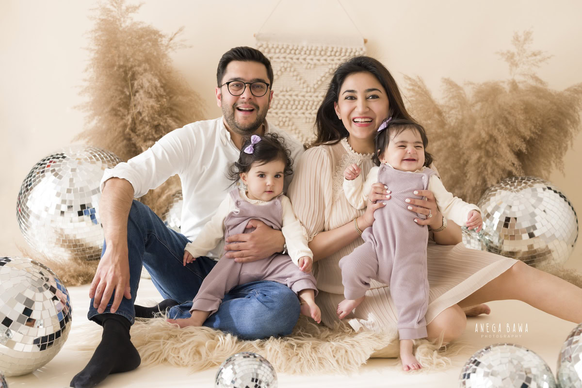 1-year-old cute twins seated on a beige rug with mom and dad, amidst bushes and disco balls on a beige backdrop, family photoshoot by Anega Bawa, Gurgaon (Delhi, NCR)