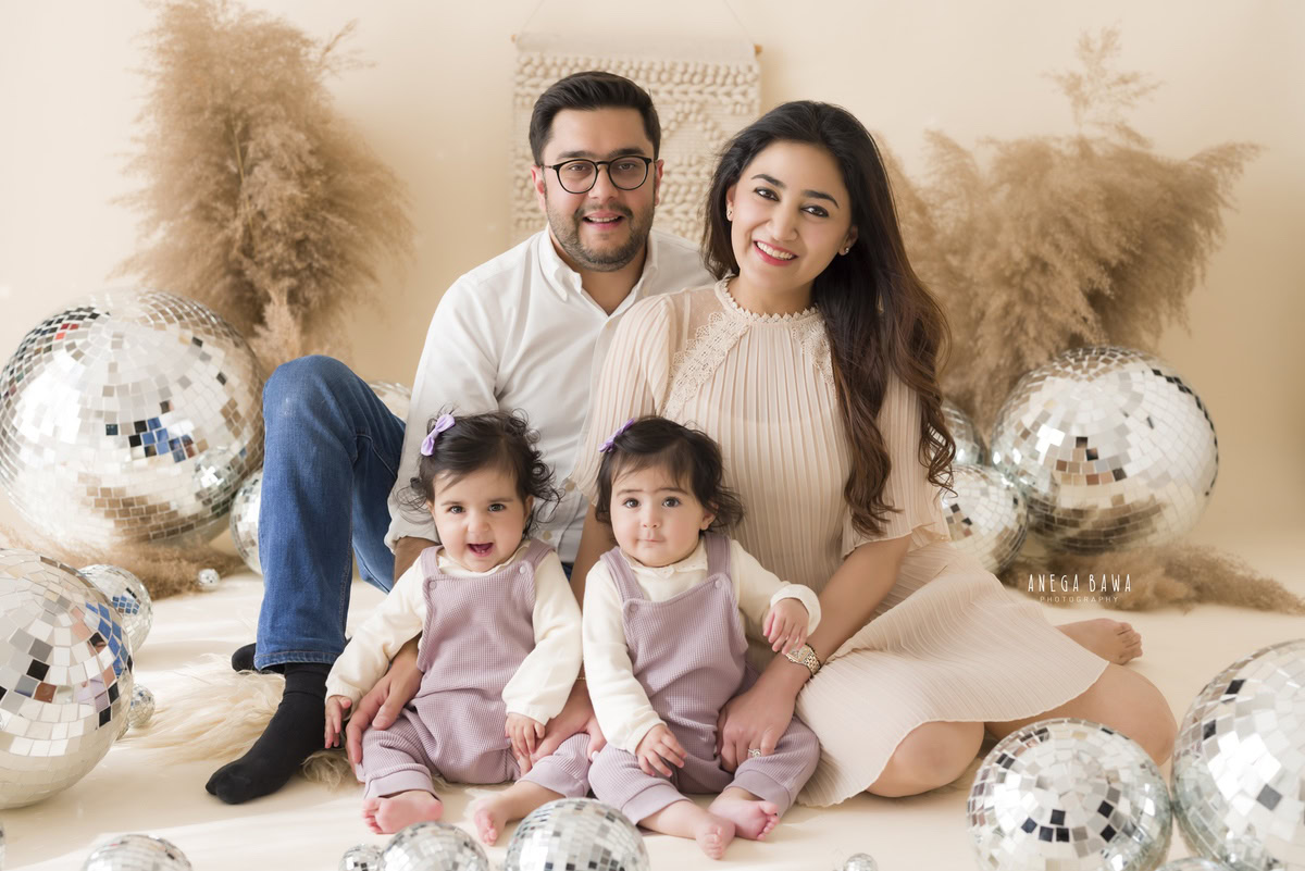 1-year-old cute twins seated on the floor with mom and dad, amidst bushes and disco balls, against a beige backdrop.