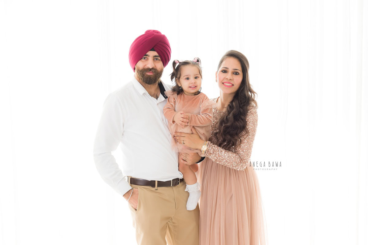 1-year-old girl in a beige dress posing cutely with mom and dad against a white backdrop, captured by Anega Bawa, family photographer in Gurgaon (Delhi NCR).