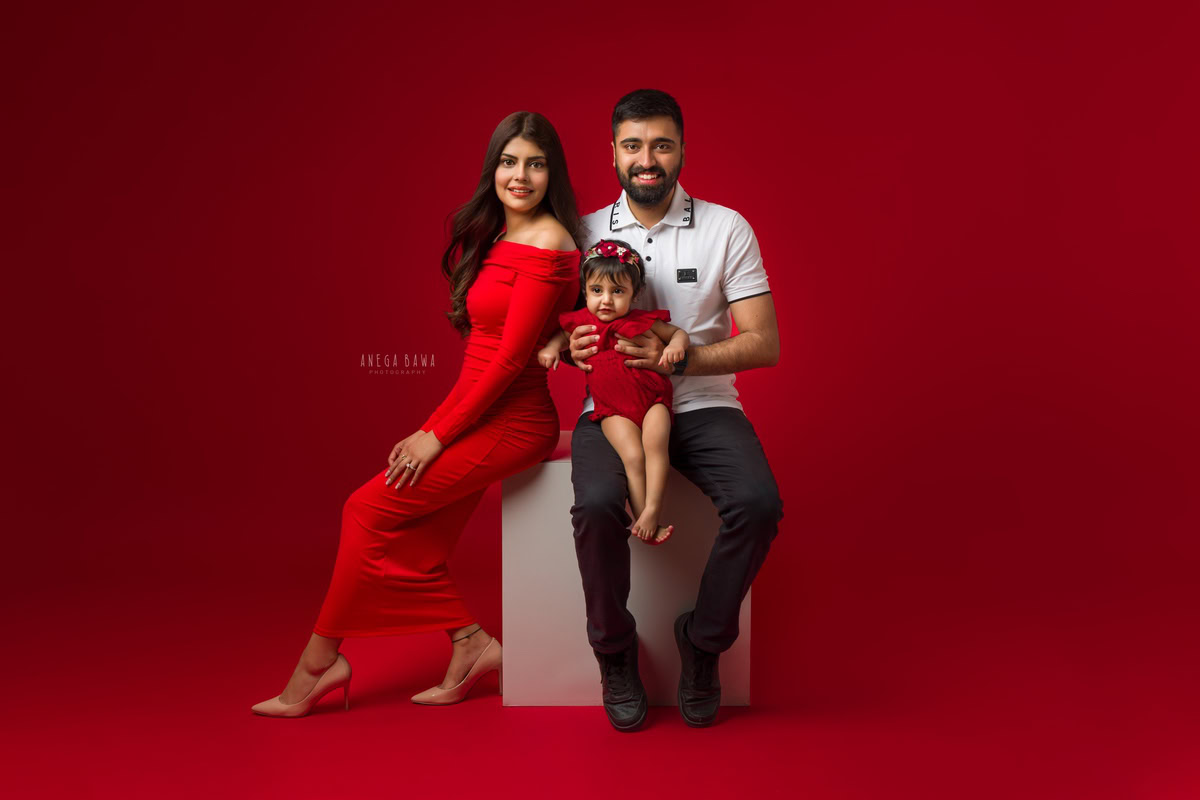 1-year-old girl in a cute red dress and red headband, posing with mom and dad against a red backdrop, captured by Anega Bawa Family Photographer Gurgaon (Delhi NCR).