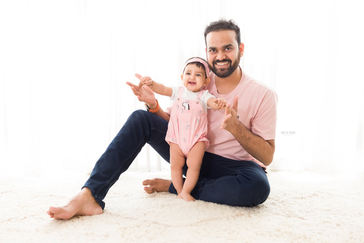 1-year-old girl holding dad's hands while seated on the floor against a serene white backdrop.