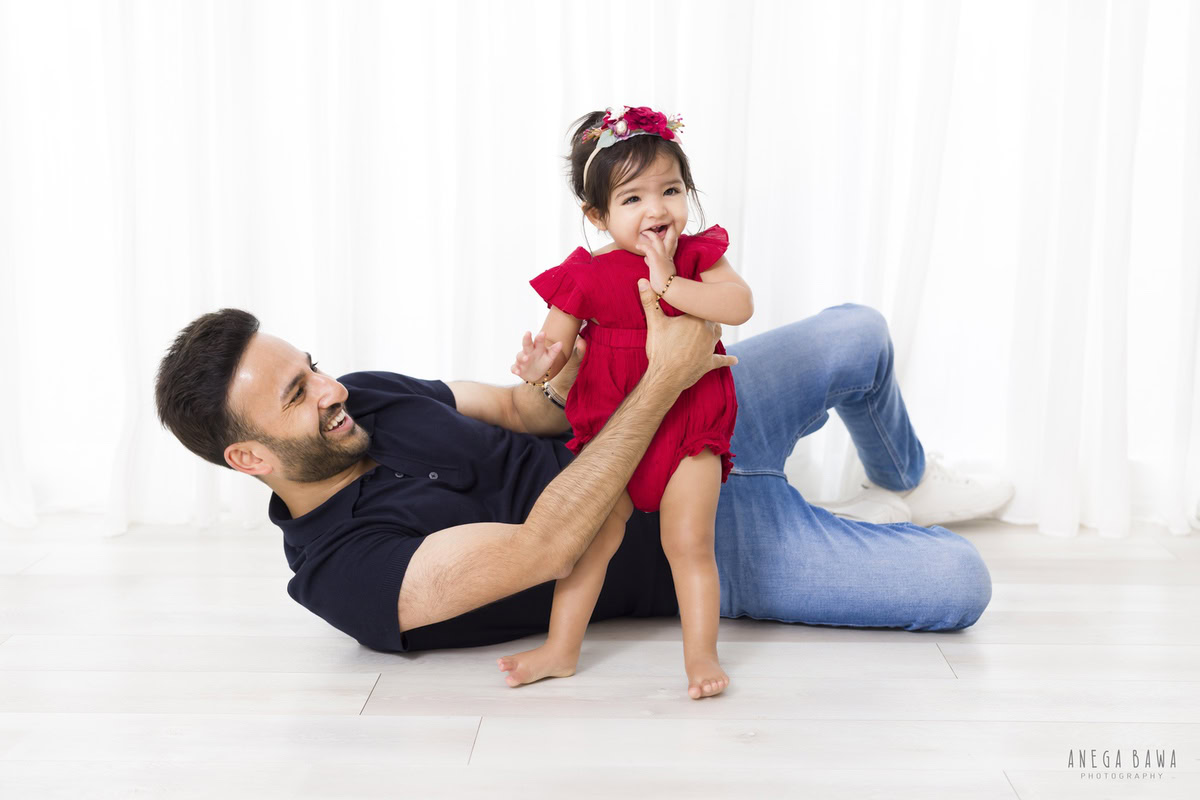 1-year-old girl in an adorable red dress poses with her dad lying on the floor against a serene white backdrop, skillfully captured by Anega Bawa, a family photographer in Gurgaon (Delhi, NCR).