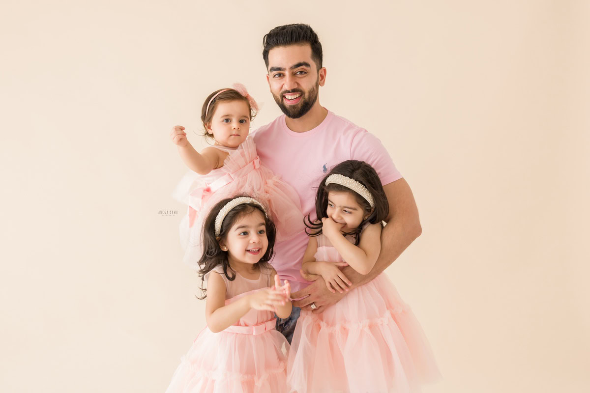 1-year-old girl in dad's arms, posing with twin girls, all wearing pastel pink against a beige backdrop, family photoshoot by Anega Bawa, Gurgaon (Delhi, NCR)