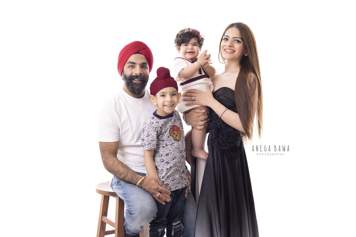 1-year-old girl in mom's arms, with her elder brother and dad in a family pose against a white backdrop, captured by Anega Bawa Family Photographer Gurgaon (Delhi NCR).