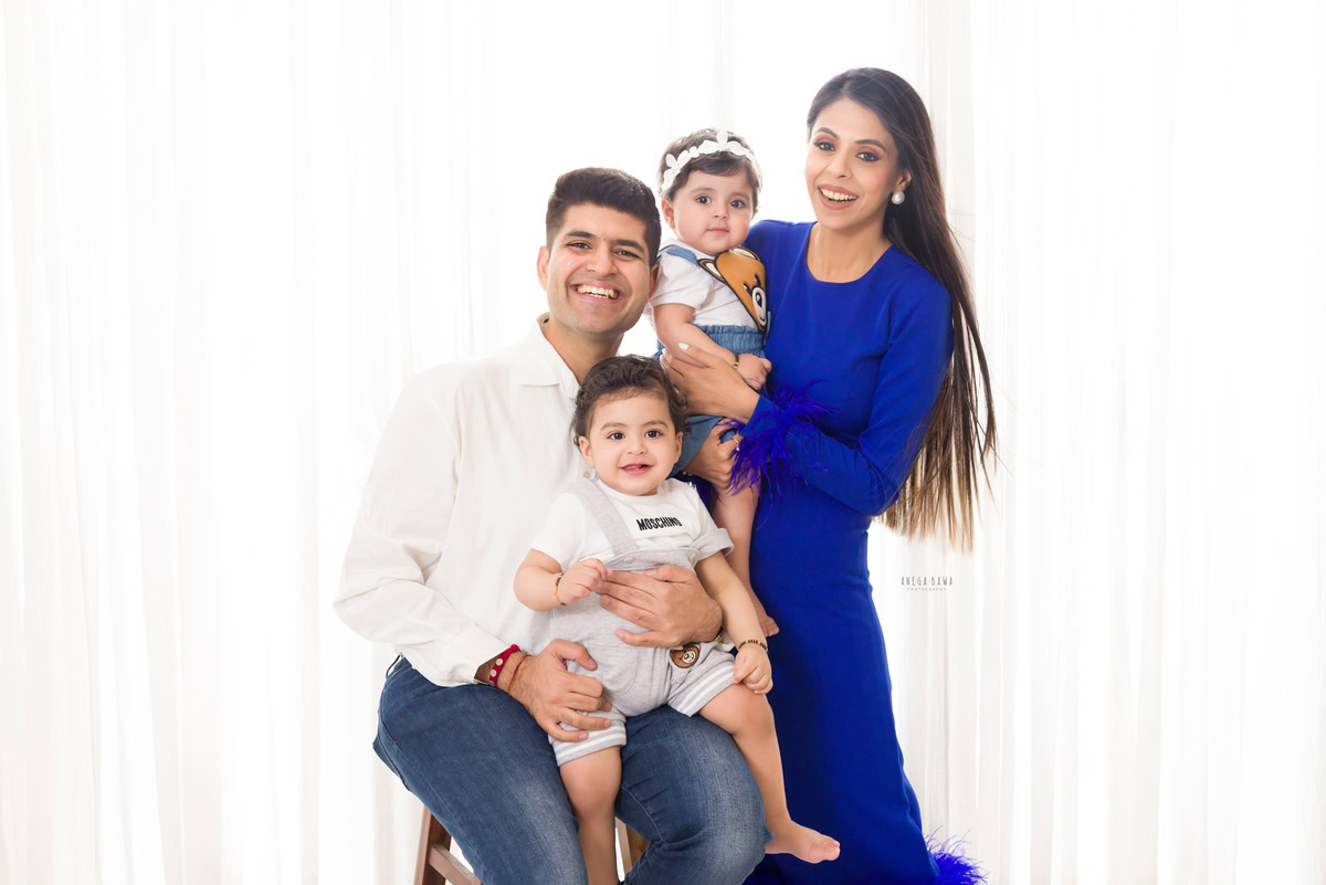 1-year-old girl in mom's arms, posing with dad and elder brother against a white backdrop, captured by Anega Bawa, family photographer in Gurgaon (Delhi NCR).