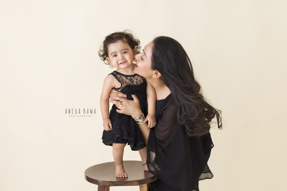 Here's the alt-text for the image based on the filename provided: 1-year-old girl in a kissing pose with mom against a beige backdrop, captured by Anega Bawa, family photographer in Gurgaon (Delhi NCR).