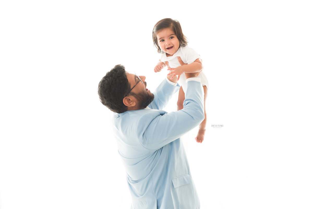 1-year-old girl joyfully lifted in her dad's arms against a bright white backdrop, showcasing a delightful and candid moment captured by Anega Bawa Photography for a family photoshoot in Gurgaon, Delhi NCR.