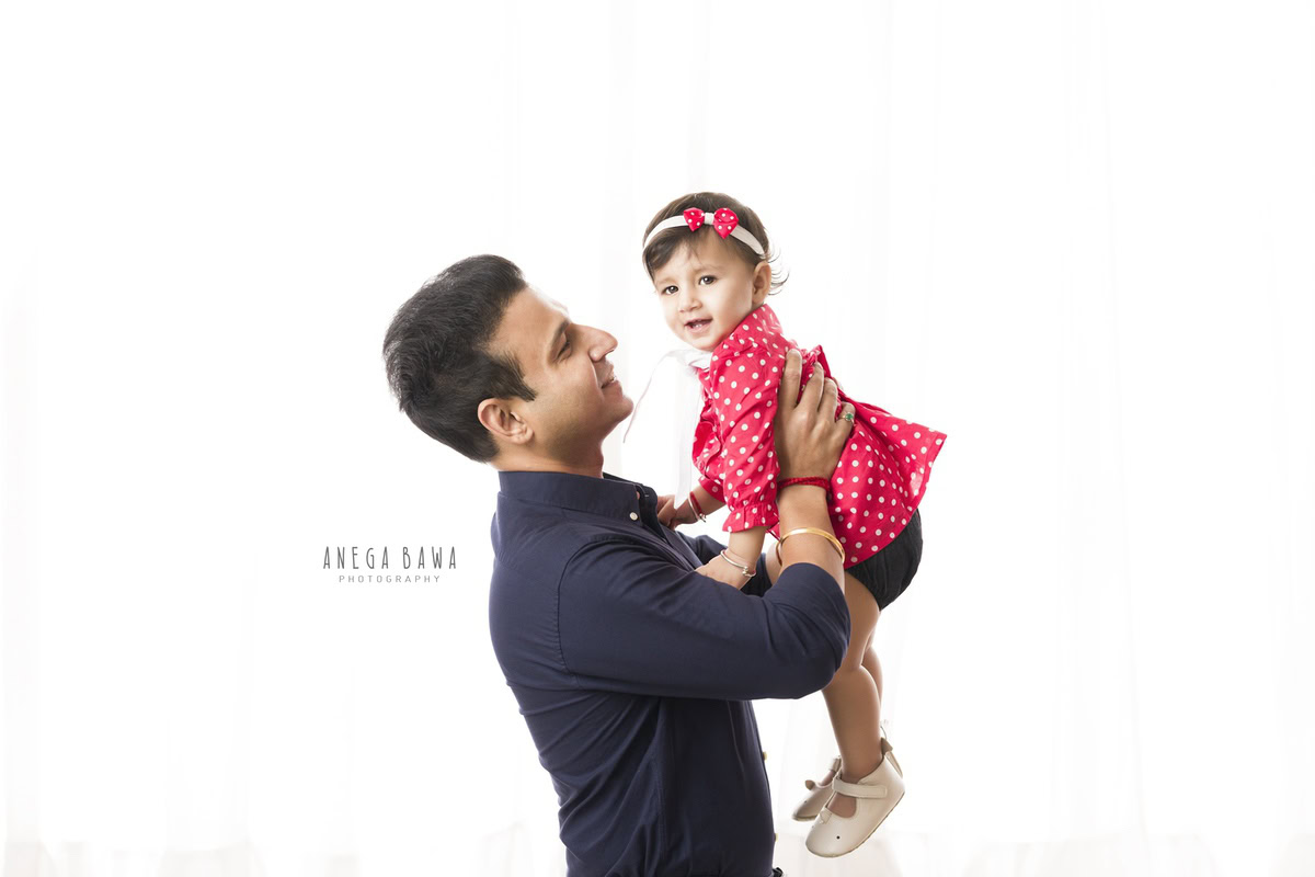 1-year-old girl lifted in dad's arms against a white backdrop, captured by Anega Bawa, family photographer in Gurgaon (Delhi NCR).
