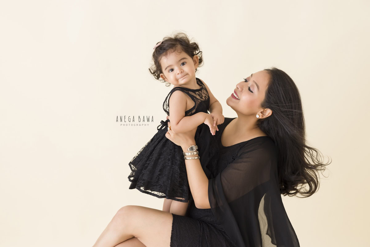 1-year-old girl lifted in mom's arms, both wearing black, against a beige backdrop, captured by Anega Bawa Family Photographer Gurgaon (Delhi NCR).