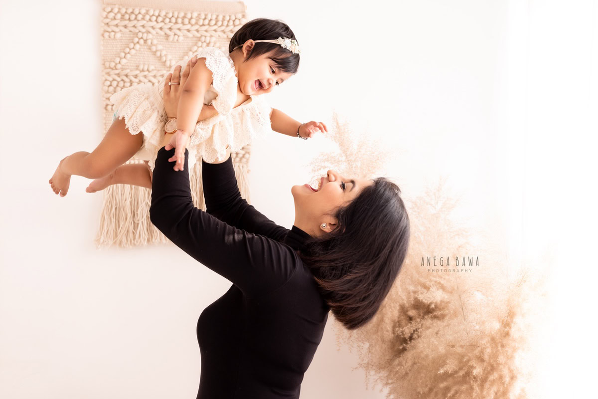 1-year-old girl lifted in mom's arms against a white backdrop, captured by Anega Bawa Family Photographer Gurgaon (Delhi NCR).