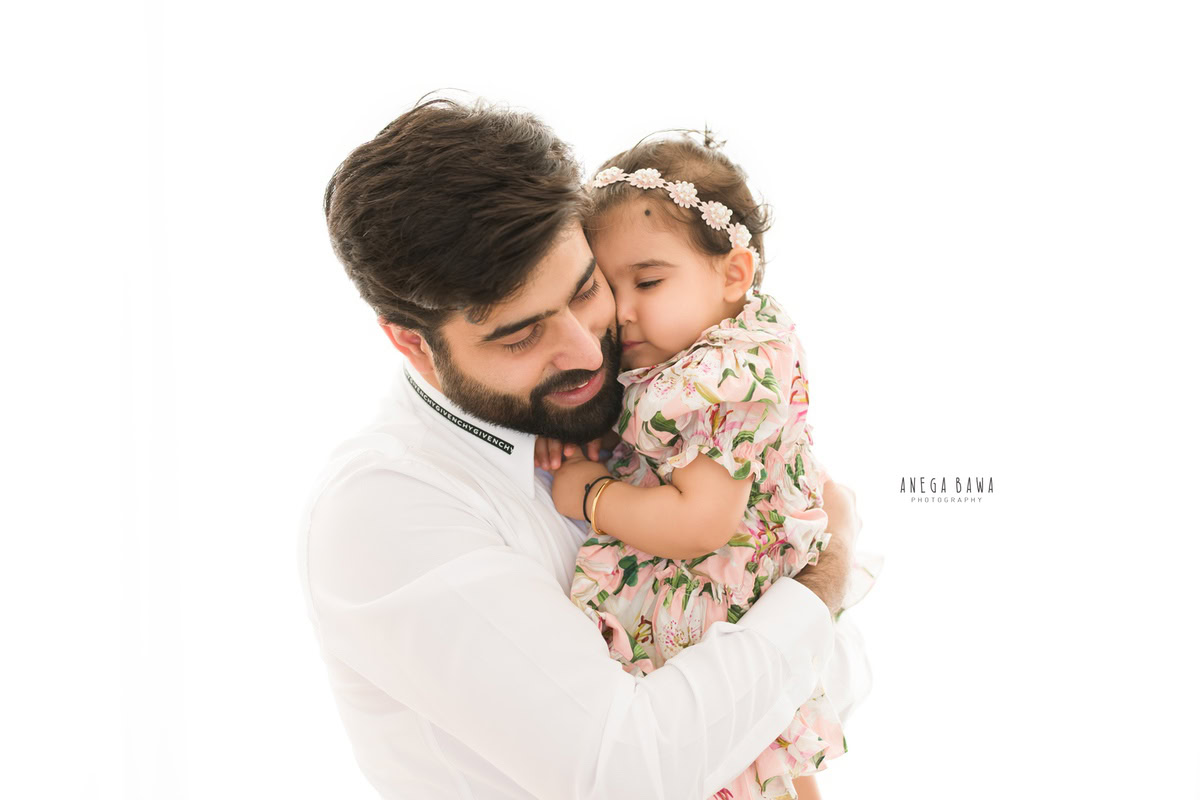 1-year-old girl lying on dad's shoulder, wearing a cute headband, against a white backdrop, family photoshoot by Anega Bawa, Gurgaon (Delhi, NCR)