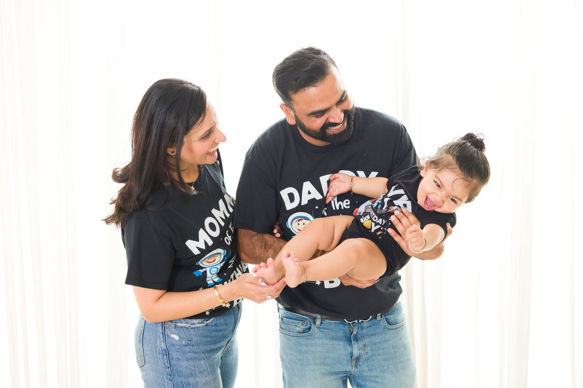 1-year-old girl playing joyfully with mom and dad in a cute, smiling pose against a white backdrop, captured by Anega Bawa Photography for a family photoshoot in Gurgaon, Delhi NCR.