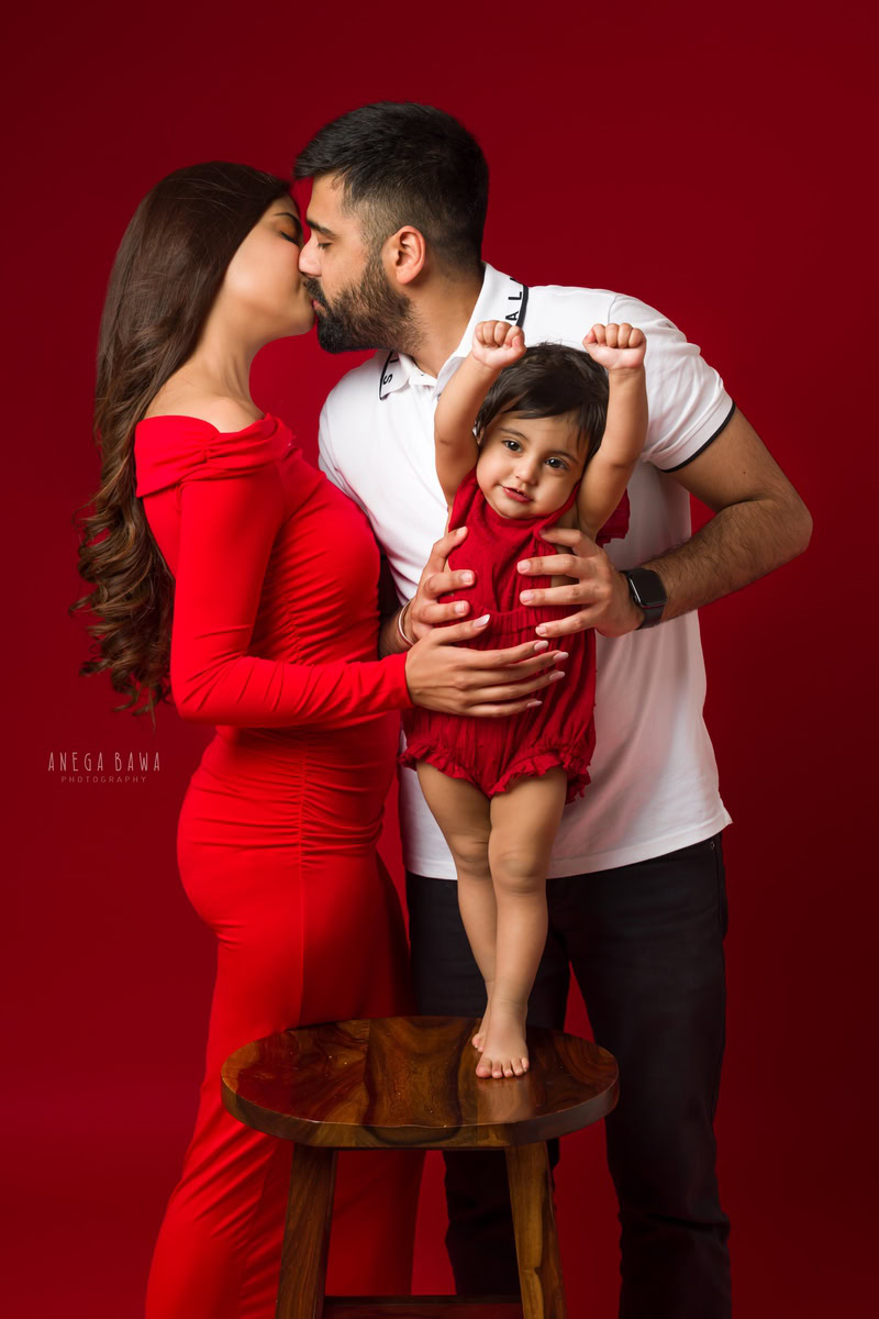 1-year-old girl playing with mom and dad against a red backdrop, capturing a tender moment with mom and dad in a kissing pose. Photographed by Anega Bawa Photography for a family photoshoot in Gurgaon (Delhi NCR).