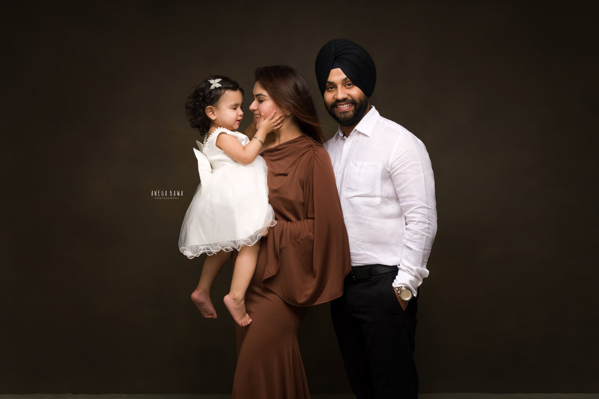 1-year-old girl playing with mom, posing with dad against a brown backdrop, captured beautifully by Anega Bawa Photography for a family photoshoot in Gurgaon (Delhi NCR).