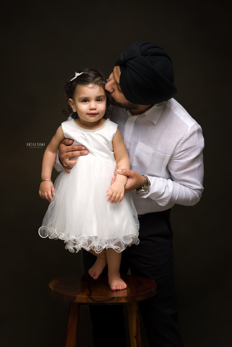 1-year-old girl poses with dad in a heartwarming kissing pose against a black backdrop, captured by Anega Bawa Photography for a family photoshoot in Gurgaon (Delhi NCR).