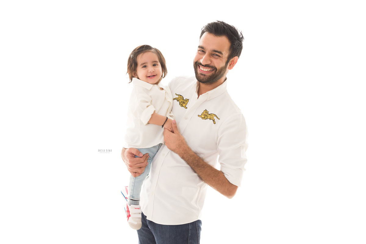 1-year-old girl poses with dad in a cute smiling pose against a white backdrop, captured by Anega Bawa Photography for a family photoshoot in Gurgaon (Delhi NCR).