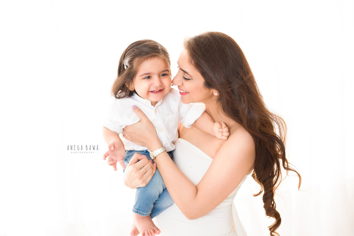 1-year-old girl poses with mom in a cute smiling pose against a white backdrop, captured by Anega Bawa Photography for a family photoshoot in Gurgaon (Delhi NCR).