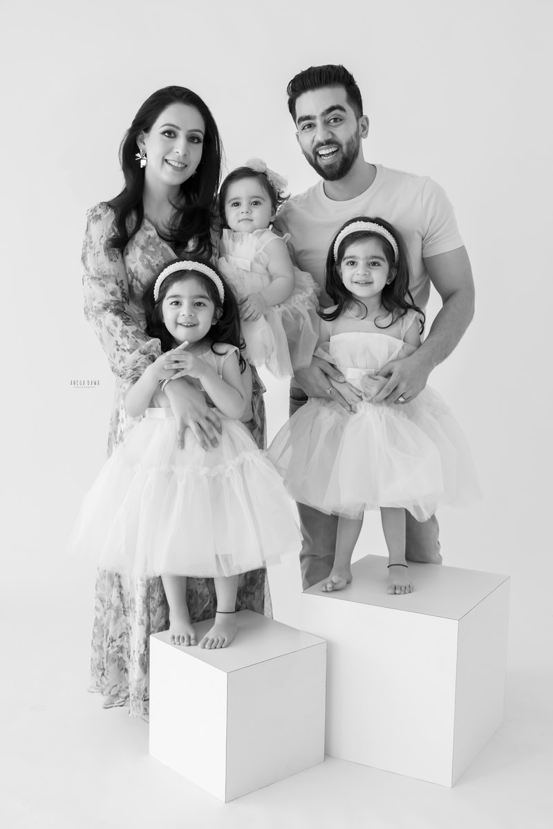 1-year-old girl posing with mom, dad, and twin sisters in a family pose against a white backdrop, captured by Anega Bawa Family Photographer Gurgaon (Delhi NCR) in black and white.