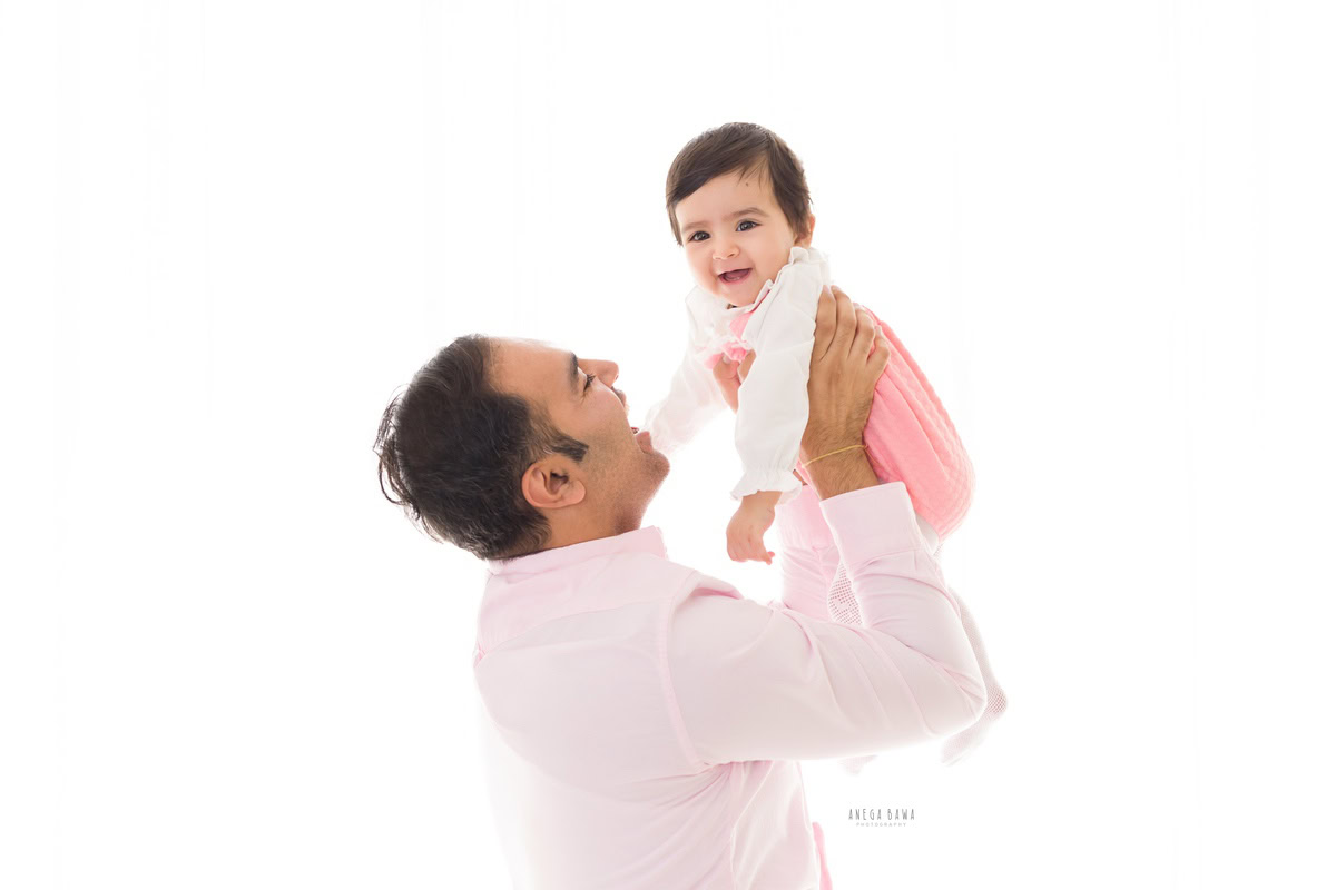 1-year-old girl posing cute in dad's arms against a serene white backdrop, photographed by Anega Bawa in Gurgaon (Delhi NCR).