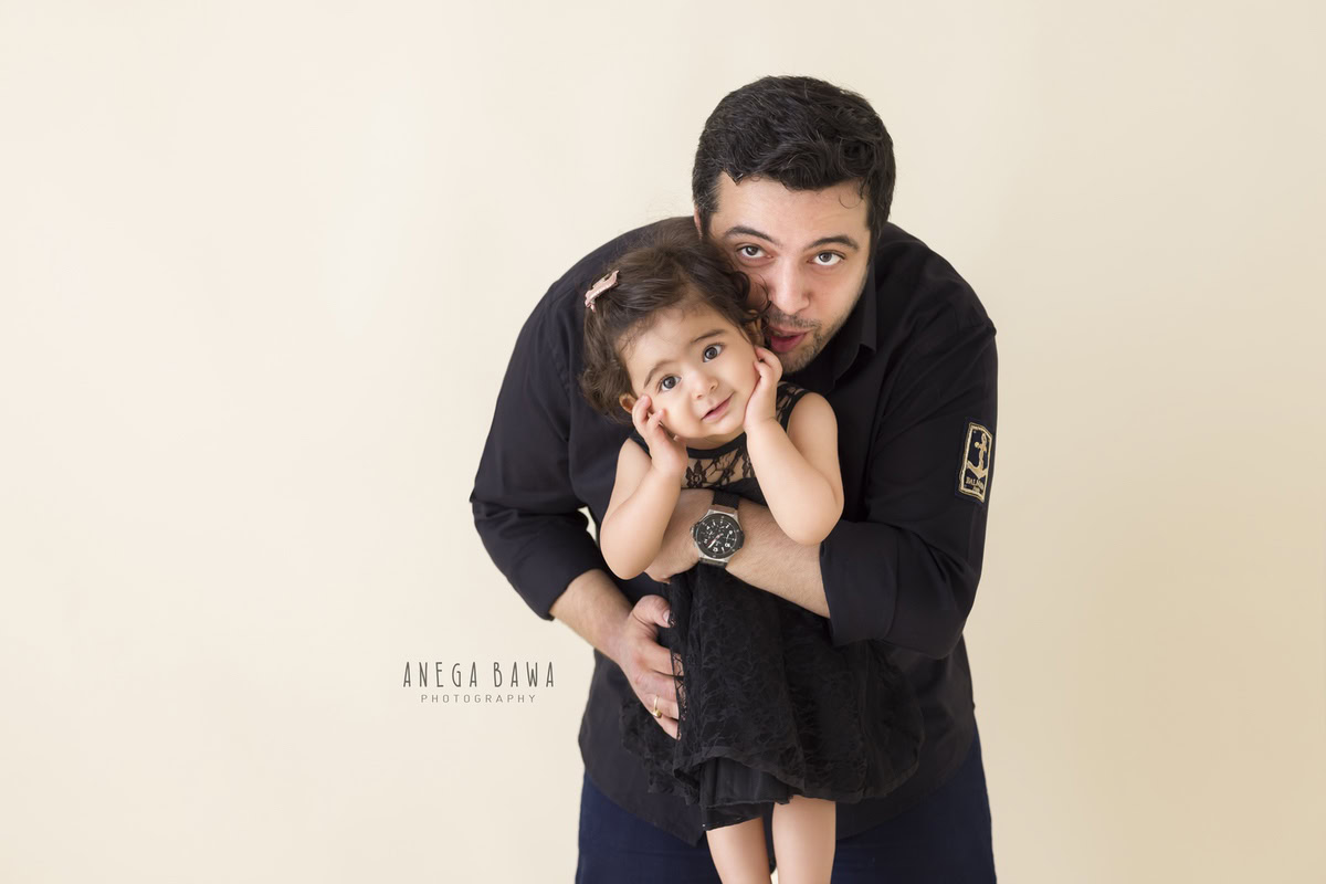 1-year-old girl posing cutely with dad against a beige backdrop, captured by Anega Bawa Family Photographer Gurgaon (Delhi NCR).