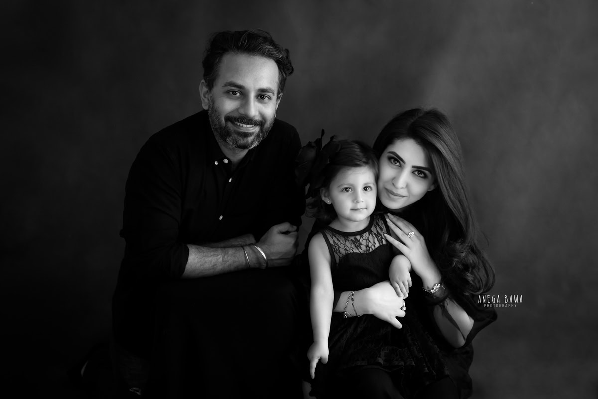 1-year-old girl posing cutely with mom and dad, all wearing black, against a black and white backdrop, captured by Anega Bawa Family Photographer Gurgaon (Delhi NCR).