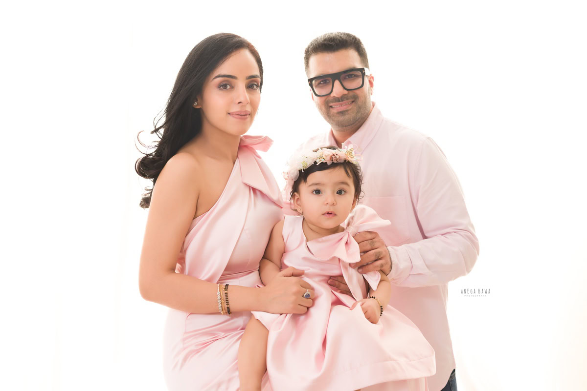 1-year-old girl posing cutely with mom and dad, all wearing pastel pink, against a white backdrop, captured by Anega Bawa Family Photographer Gurgaon (Delhi NCR).