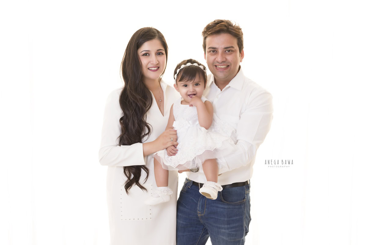 1-year-old girl posing cutely with mom and dad, all wearing white, against a white backdrop, family photoshoot by Anega Bawa, Gurgaon (Delhi, NCR)