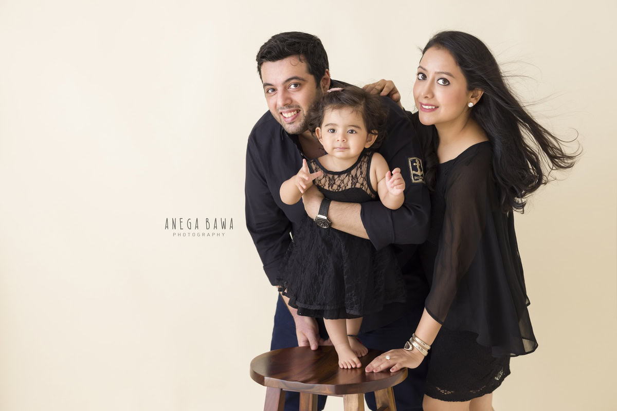 1-year-old girl posing cutely with mom and dad against a beige backdrop, captured by Anega Bawa, family photographer in Gurgaon (Delhi NCR).