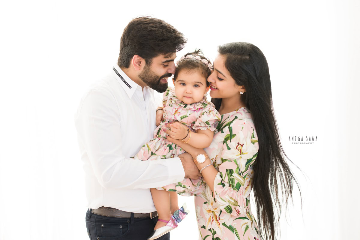1-year-old girl in a floral dress posing cutely with mom and dad against a white backdrop, captured by Anega Bawa, family photographer in Gurgaon (Delhi NCR).