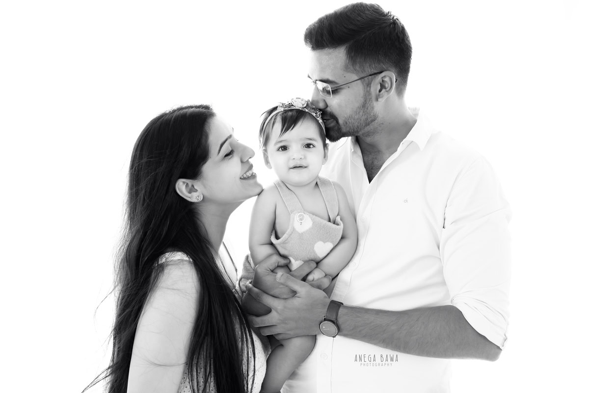 Here's the alt-text for the image based on the filename provided: 1-year-old girl posing cutely with mom and dad in a kissing pose against a white backdrop, captured by Anega Bawa, family photographer in Gurgaon (Delhi NCR).