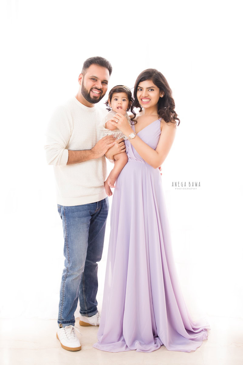 1-year-old girl posing cutely with mom and dad while standing on the floor against a white backdrop, captured by Anega Bawa, family photographer in Gurgaon (Delhi NCR).